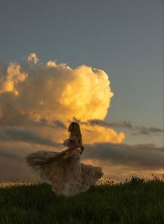 a woman in a long dress standing on top of a lush green field under a cloudy sky