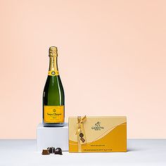 a bottle of champagne next to a box of chocolates on a white table with a pink background