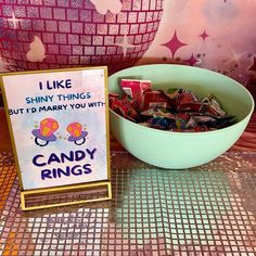 a bowl of candy sits next to a sign that says i like shiny things but i'd marry you with candy rings