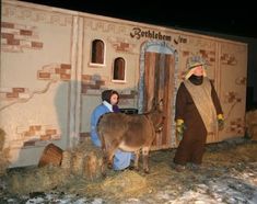 two people are standing in front of a small house with a donkey and hay bales