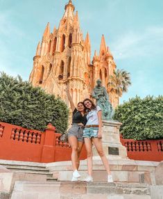 two women standing on steps in front of a large building with statues and trees around it