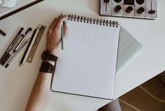 a person holding a pen and writing on a notepad next to some markers, pencils and other office supplies