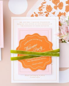 an orange and pink wedding card with a green ribbon on the front next to some flowers