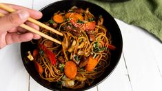 someone holding chopsticks over a bowl of noodles with meat and veggies