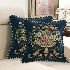 two blue pillows with flowers on them sitting on a white couch in front of a painting