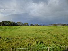 a green field with trees and mountains in the distance under a cloudy sky, viewed from a train window