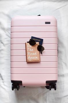 a pink suitcase with sunglasses and a passport on it sitting on a white sheet that says the road again