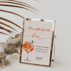 a sign with an orange drink on it next to some flowers and leaves in front of a white wall