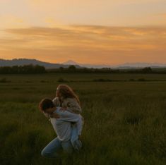 two people are hugging in the middle of a field with mountains in the background at sunset