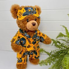 a brown teddy bear sitting next to a green plant and wearing a yellow shirt with bears on it