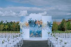 an outdoor ceremony setup with white chairs and flowers on the wall, in front of trees