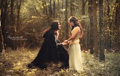 two women standing in the woods with one holding onto another's hand and looking at each other