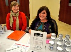 two women are sitting at a table with jars and papers on it, smiling for the camera