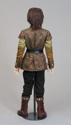 the back view of a doll with brown hair and black pants, standing on a stand