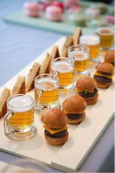 mini burgers and beer are lined up on a long tray with glasses of beer