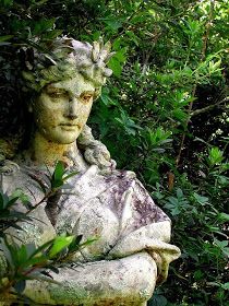 an old statue in the middle of some bushes and trees with its arms folded out