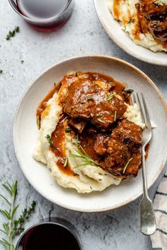 two plates with meat and mashed potatoes on them next to glasses of red wine