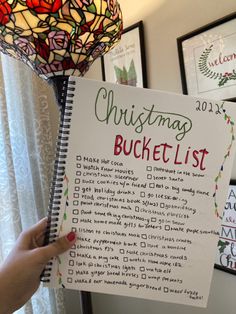 a hand holding a christmas bucket list in front of a lamp with pictures on the wall behind it