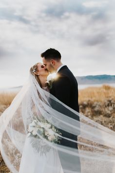 a bride and groom kissing in the middle of an open field with their veil blowing in the wind