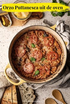 a pizza in a pan with basil, tomatoes and olives on the side next to some bread