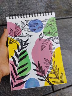 a hand holding a notebook with colorful flowers and leaves painted on the cover, while it's being held by someone