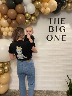 a woman holding a baby in front of a wall with balloons and the words the big one on it