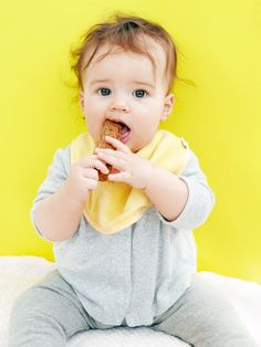 a baby sitting on the floor eating something with his mouth wide open and wearing a yellow bib