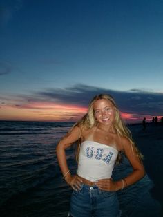 a beautiful young woman standing on top of a beach next to the ocean at sunset