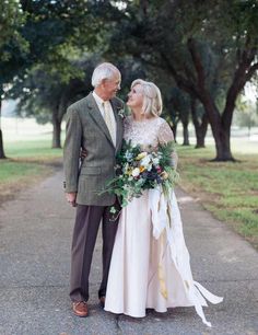 an older man and woman standing next to each other on a road with trees in the background