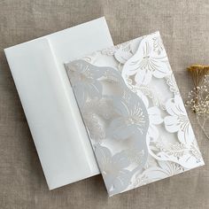 an open white wedding card on a table next to some flowers and a dried plant