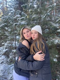 two girls hugging each other in front of snow - covered trees and evergreens on a sunny day