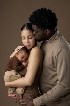 a man and woman holding a baby while standing next to each other in front of a brown background