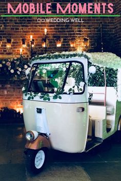 a small white and green vehicle with flowers on the back is parked in front of a brick wall