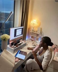 a woman sitting at a desk with a laptop and computer monitor on it, in front of a window