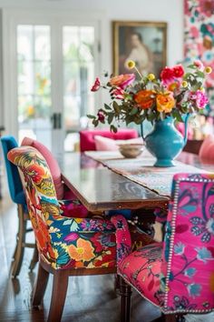 a dining room table with colorful chairs and flowers in a vase on the top of it