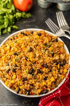 mexican rice and beans in a white bowl on a black surface with silverware, tomatoes, and lettuce