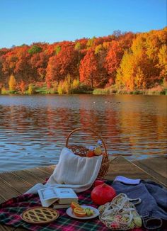 an open book and some food on a picnic blanket by the water with autumn trees in the background