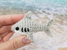 a hand holding a small crocheted toy shark on the beach with waves in the background