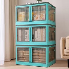 a teal colored storage unit with glass doors and shelves filled with books in front of a beige chair