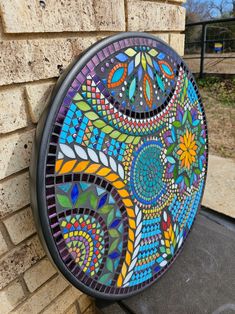 a colorful stained glass plate hanging on the side of a brick wall next to a black trash can