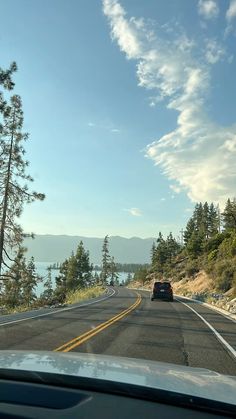 a car driving down the road next to some pine trees and water with mountains in the background