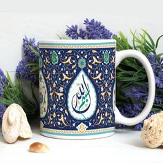 a blue and white coffee mug with an arabic calligraphy on it next to purple flowers