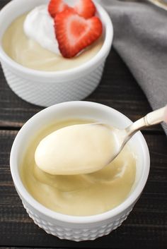 two white bowls filled with pudding and strawberries on top of a wooden table next to a spoon