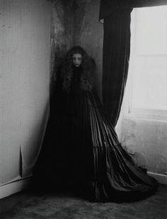 a woman in a long black dress standing next to a window with curtains on it