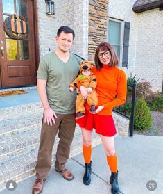 a man and woman standing in front of a house holding a small dog wearing an orange outfit