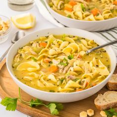 two bowls of chicken noodle soup on a cutting board with bread and parsley