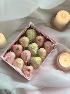 a pink box filled with chocolate covered strawberries next to two lit candles on a white cloth