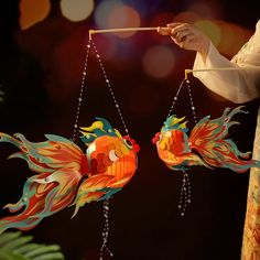 two colorful birds hanging from strings in front of a person holding a stick and wearing a white shirt