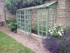 a small green house sitting in the middle of a garden next to a stone wall