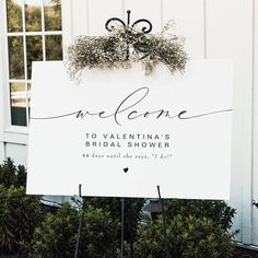 a welcome sign in front of a white building with potted plants and greenery